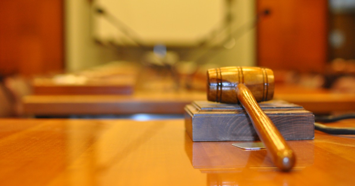 A wooden gavel rests on a block atop a polished table in a courtroom. The background is softly blurred.